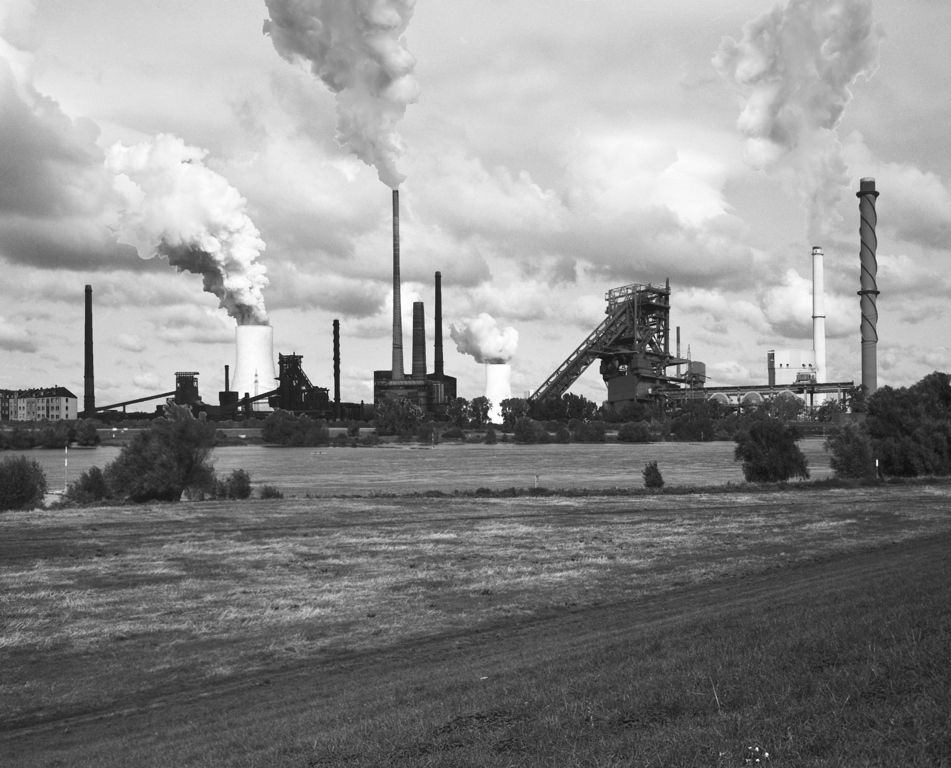 Ein Schwarz-Weiß-Bild zeigt ein industrielles Panorama mit rauchenden Schornsteinen und einer Stahlproduktion im Hintergrund. Im Vordergrund erstrecken sich eine weite Wiese und ein Fluß.