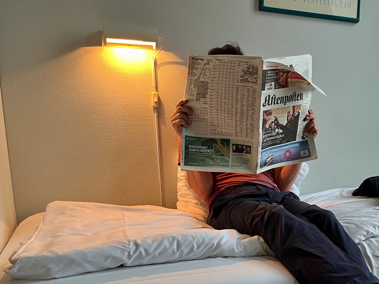 Eine Person sitzt auf einem Bett und liest die Zeitung "Aftenposten". Eine Lampe leuchtet über dem Bett.
